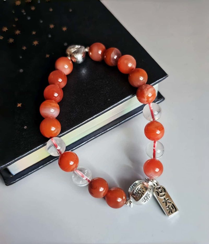 AB-ACT-01-01 Damenarmband aus rotem Achat Kugeln Bergkristall fein poliert versilberten Details mit chinesischen Schriftzeichen. Flache Ansicht mit einem Buch