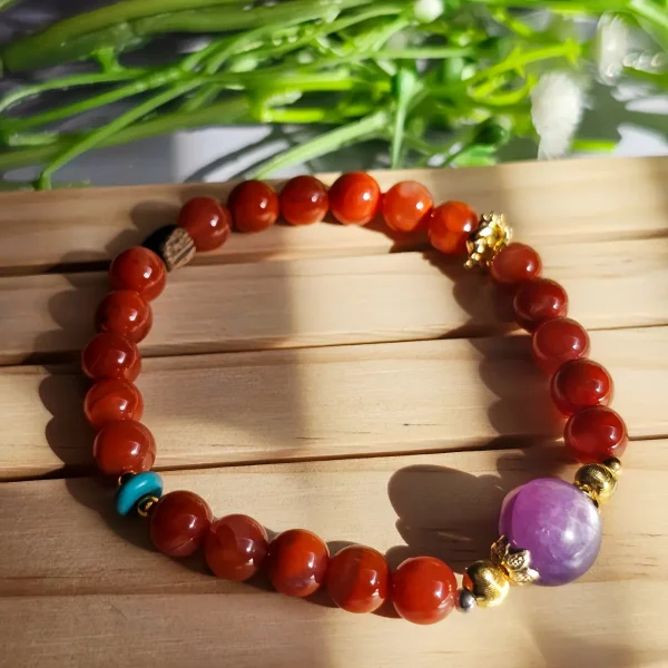 Achat-AB02-01 ein rotes Damenarmband mit Roter Achat Naturstein und violetter Lepidolith-Zentralperle, goldfarbenen Zierelementen und kleinen türkisfarbenen Akzentperlen auf ein Holzplatte, vor eine Blumenhintergrund