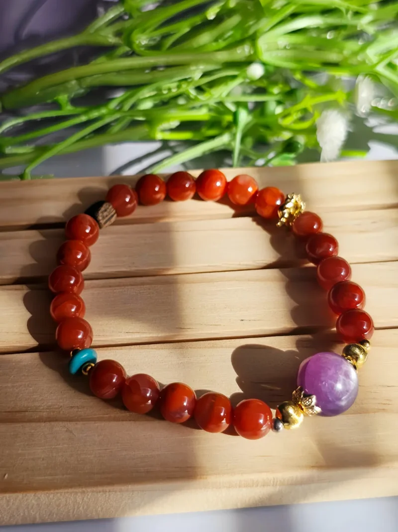 Achat-AB02-01 ein rotes Damenarmband mit Roter Achat Naturstein und violetter Lepidolith-Zentralperle, goldfarbenen Zierelementen und kleinen türkisfarbenen Akzentperlen auf ein Holzplatte, vor eine Blumenhintergrund