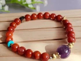 Achat-AB02-02 ein rotes Damenarmband mit Roter Achat Naturstein und violetter Lepidolith-Zentralperle, goldfarbenen Zierelementen und kleinen türkisfarbenen Akzentperlen auf einem Holzplatte, vor einer Blumenhintergrund