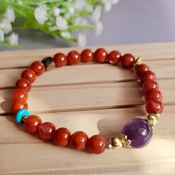 Achat-AB02-02 ein rotes Damenarmband mit Roter Achat Naturstein und violetter Lepidolith-Zentralperle, goldfarbenen Zierelementen und kleinen türkisfarbenen Akzentperlen auf einem Holzplatte, vor einer Blumenhintergrund