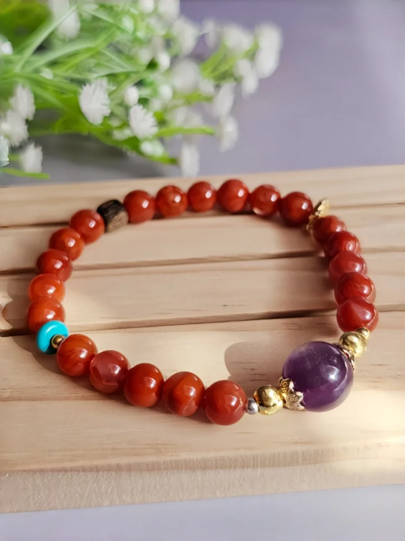 Achat-AB02-02 ein rotes Damenarmband mit Roter Achat Naturstein und violetter Lepidolith-Zentralperle, goldfarbenen Zierelementen und kleinen türkisfarbenen Akzentperlen auf einem Holzplatte, vor einer Blumenhintergrund
