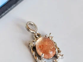 Das Bild zeigt einen detailreichen Anhänger mit einem ovalen, orangefarbenen Sonnenstein Edelstein , der von einem silberfarbenen, verzierten Rahmen mit kleinen funkelnden Zirkonia Steinen umgeben ist. Der Anhänger liegt auf einer hellen Oberfläche neben einer Illustration von orangefarbenen Früchten an einem Zweig.