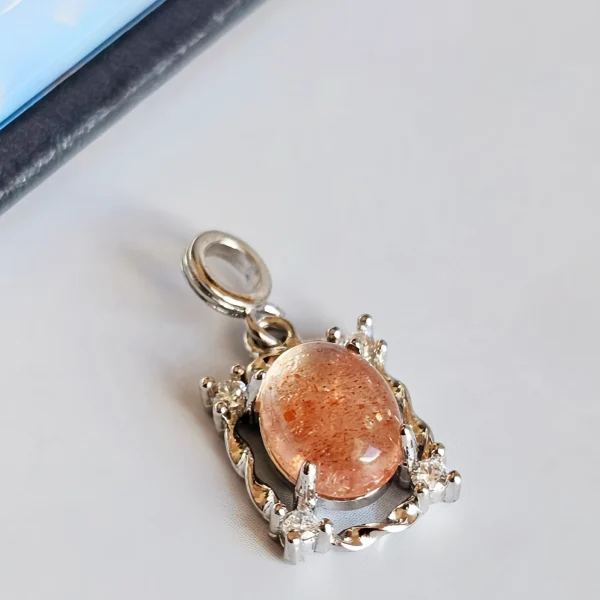 Sonnestein Naturstein Anhaenger Eleganter Schmuck Das Bild zeigt einen detailreichen Anhänger mit einem ovalen, orangefarbenen Sonnenstein Edelstein , der von einem silberfarbenen, verzierten Rahmen mit kleinen funkelnden Zirkonia Steinen umgeben ist. Der Anhänger liegt auf einer hellen Oberfläche neben einer Illustration von orangefarbenen Früchten an einem Zweig.