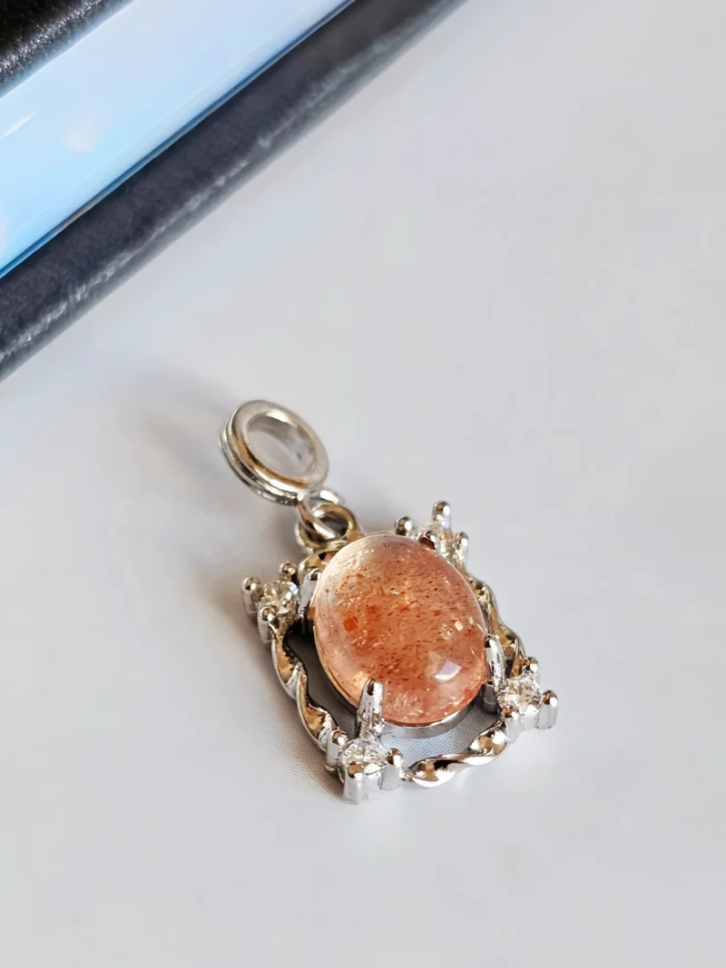 Sonnestein Naturstein Anhaenger Eleganter Schmuck Das Bild zeigt einen detailreichen Anhänger mit einem ovalen, orangefarbenen Sonnenstein Edelstein , der von einem silberfarbenen, verzierten Rahmen mit kleinen funkelnden Zirkonia Steinen umgeben ist. Der Anhänger liegt auf einer hellen Oberfläche neben einer Illustration von orangefarbenen Früchten an einem Zweig.