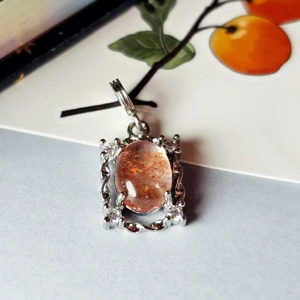 HK-TDS-08-05 Das Bild zeigt einen detailreichen Anhänger mit einem ovalen, orangefarbenen Sonnenstein, der von einem silberfarbenen, verzierten Rahmen mit kleinen funkelnden Zirkonia Steinen umgeben ist. Der Anhänger liegt auf einer hellen Oberfläche neben einer Illustration von orangefarbenen Früchten an einem Zweig.
