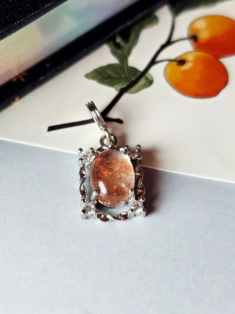 HK-TDS-08-05 Das Bild zeigt einen detailreichen Anhänger mit einem ovalen, orangefarbenen Sonnenstein, der von einem silberfarbenen, verzierten Rahmen mit kleinen funkelnden Zirkonia Steinen umgeben ist. Der Anhänger liegt auf einer hellen Oberfläche neben einer Illustration von orangefarbenen Früchten an einem Zweig.