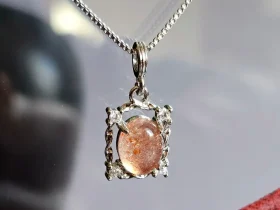 Sonnestein Naturstein Anhaenger Eleganter Schmuck Das Bild zeigt einer detailreiche Halskette mit einem ovalen, orangefarbenen Sonnenstein, die von einem silberfarbenen, verzierten Rahmen mit kleinen funkelnden Zirkonia Steinen umgeben ist. Die Halskette liegt auf einer hellen Oberfläche neben einer Illustration von orangefarbenen Früchten an einem Zweig.