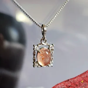 Das Bild zeigt einer detailreiche Halskette mit einem ovalen, orangefarbenen Sonnenstein, die von einem silberfarbenen, verzierten Rahmen mit kleinen funkelnden Zirkonia Steinen umgeben ist. Die Halskette liegt auf einer hellen Oberfläche neben einer Illustration von orangefarbenen Früchten an einem Zweig.