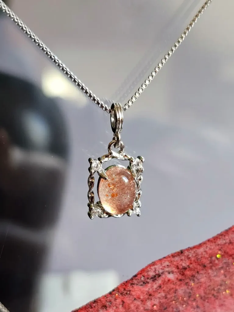 Das Bild zeigt einer detailreiche Halskette mit einem ovalen, orangefarbenen Sonnenstein, die von einem silberfarbenen, verzierten Rahmen mit kleinen funkelnden Zirkonia Steinen umgeben ist. Die Halskette liegt auf einer hellen Oberfläche neben einer Illustration von orangefarbenen Früchten an einem Zweig.
