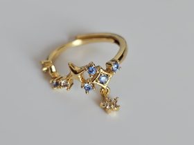Goldener Planeten-Ring mit Stern-Charms Ein goldfarbener, offener Ring mit einem Design aus Sternen und blauen sowie klaren Zirkonia-Steinen, von dem ein kleiner, mit Steinen besetzter Sternanhänger herabhängt.