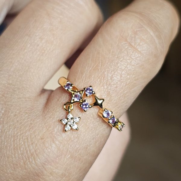 Ein Hand trägt einen goldfarbener, offener Ring mit einem Design aus Sternen und Lila sowie klaren Zirkonia-Steinen, von dem ein kleiner, mit Steinen besetzter Sternanhänger herabhängt.