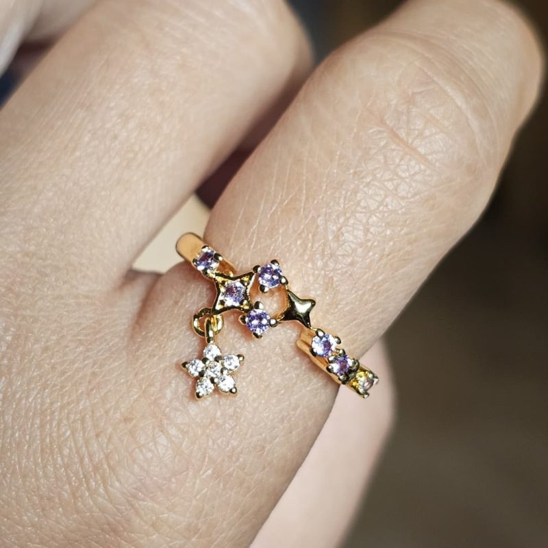 Ein Hand trägt einen goldfarbener, offener Ring mit einem Design aus Sternen und Lila sowie klaren Zirkonia-Steinen, von dem ein kleiner, mit Steinen besetzter Sternanhänger herabhängt.