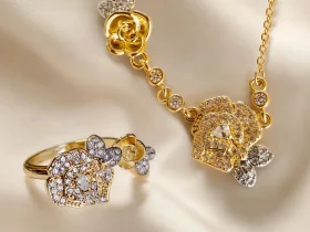 SET-TDG-01-01 Nahaufnahme einer goldfarbenen Geschenke Schmuckset-Halskette und Damenring. Der Set zeigt zwei stilisierte Rosenblüten: Eine Blüte ist glatt und poliert, die andere ist dicht mit kleinen, funkelnden Kristallen (Pavé-Optik) besetzt und hat einen größeren Kristall in der Mitte. Die Kette verbindet die Elemente mit kleinen, runden Kristallsteinen.