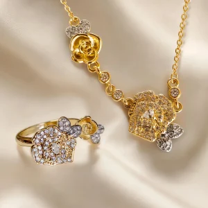 Nahaufnahme einer goldfarbenen Geschenke Schmuckset-Halskette und Damenring. Der Set zeigt zwei stilisierte Rosenblüten: Eine Blüte ist glatt und poliert, die andere ist dicht mit kleinen, funkelnden Kristallen (Pavé-Optik) besetzt und hat einen größeren Kristall in der Mitte. Die Kette verbindet die Elemente mit kleinen, runden Kristallsteinen.