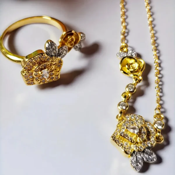 Schmuckset mit Halskette und Damenring in Rosen Design Nahaufnahme einer goldfarbenen Geschenke Schmuckset-Halskette und Damenring. Der Set zeigt zwei stilisierte Rosenblüten: Eine Blüte ist glatt und poliert, die andere ist dicht mit kleinen, funkelnden Kristallen (Pavé-Optik) besetzt und hat einen größeren Kristall in der Mitte. Die Kette verbindet die Elemente mit kleinen, runden Kristallsteinen.