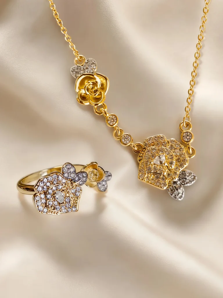 Schmuckset - Halskette und Ring Set in aufwendigem Doppel-Blüten Design Nahaufnahme einer goldfarbenen Geschenke Schmuckset-Halskette und Damenring. Der Set zeigt zwei stilisierte Rosenblüten: Eine Blüte ist glatt und poliert, die andere ist dicht mit kleinen, funkelnden Kristallen (Pavé-Optik) besetzt und hat einen größeren Kristall in der Mitte. Die Kette verbindet die Elemente mit kleinen, runden Kristallsteinen.