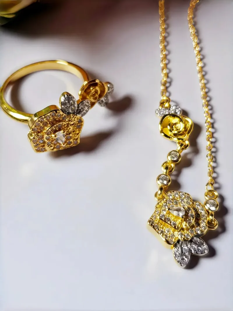 Schmuckset mit Halskette und Damenring in Rosen Design Nahaufnahme einer goldfarbenen Geschenke Schmuckset-Halskette und Damenring. Der Set zeigt zwei stilisierte Rosenblüten: Eine Blüte ist glatt und poliert, die andere ist dicht mit kleinen, funkelnden Kristallen (Pavé-Optik) besetzt und hat einen größeren Kristall in der Mitte. Die Kette verbindet die Elemente mit kleinen, runden Kristallsteinen.