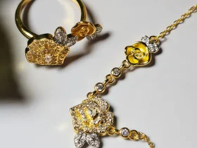 Schmuckset mit Halskette und Damenring in Rosen Design Nahaufnahme einer goldfarbenen Geschenke Schmuckset-Halskette und Damenring. Der Set zeigt zwei stilisierte Rosenblüten: Eine Blüte ist glatt und poliert, die andere ist dicht mit kleinen, funkelnden Kristallen (Pavé-Optik) besetzt und hat einen größeren Kristall in der Mitte. Die Kette verbindet die Elemente mit kleinen, runden Kristallsteinen.