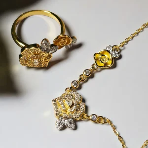 Nahaufnahme einer goldfarbenen Geschenke Schmuckset-Halskette und Damenring. Der Set zeigt zwei stilisierte Rosenblüten: Eine Blüte ist glatt und poliert, die andere ist dicht mit kleinen, funkelnden Kristallen (Pavé-Optik) besetzt und hat einen größeren Kristall in der Mitte. Die Kette verbindet die Elemente mit kleinen, runden Kristallsteinen.