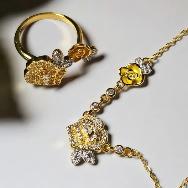 Schmuckset mit Halskette und Damenring in Rosen Design Nahaufnahme einer goldfarbenen Geschenke Schmuckset-Halskette und Damenring. Der Set zeigt zwei stilisierte Rosenblüten: Eine Blüte ist glatt und poliert, die andere ist dicht mit kleinen, funkelnden Kristallen (Pavé-Optik) besetzt und hat einen größeren Kristall in der Mitte. Die Kette verbindet die Elemente mit kleinen, runden Kristallsteinen.