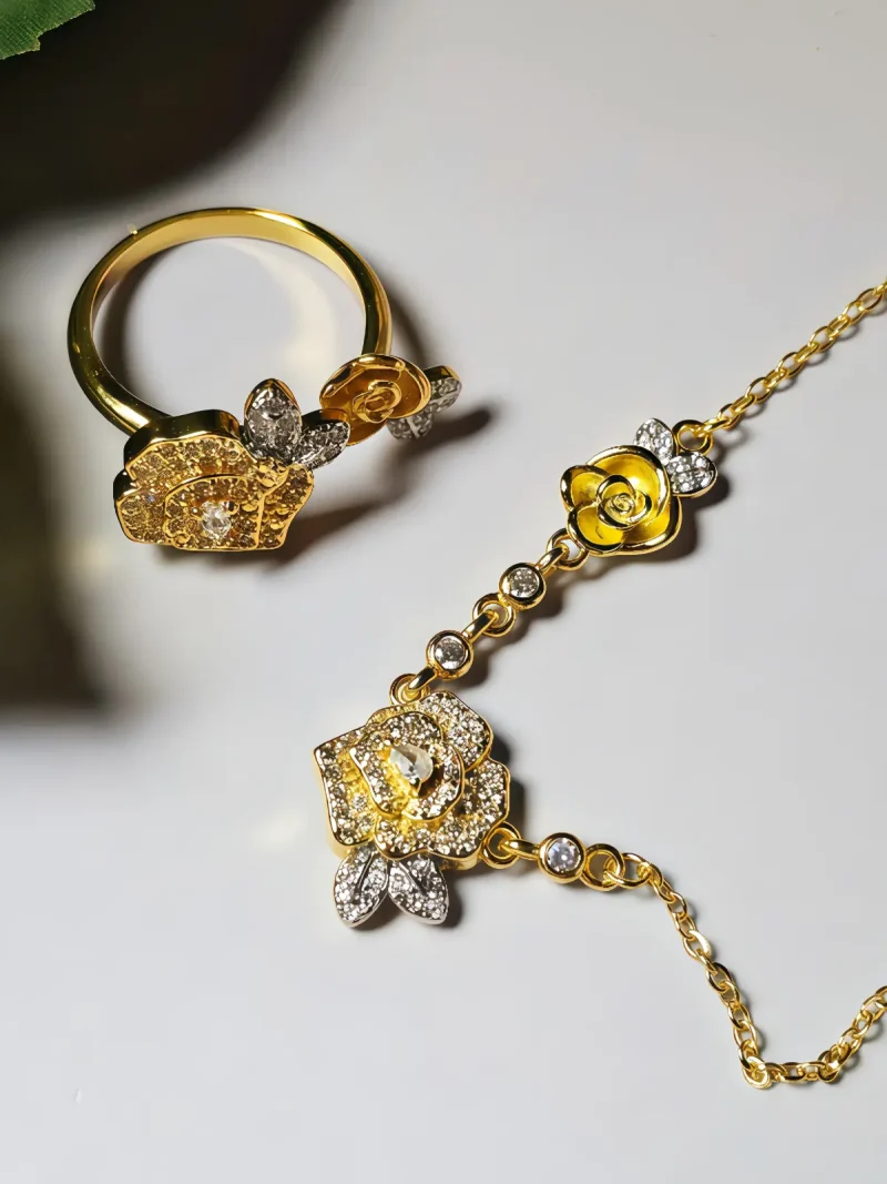 Schmuckset mit Halskette und Damenring in Rosen Design Nahaufnahme einer goldfarbenen Geschenke Schmuckset-Halskette und Damenring. Der Set zeigt zwei stilisierte Rosenblüten: Eine Blüte ist glatt und poliert, die andere ist dicht mit kleinen, funkelnden Kristallen (Pavé-Optik) besetzt und hat einen größeren Kristall in der Mitte. Die Kette verbindet die Elemente mit kleinen, runden Kristallsteinen.