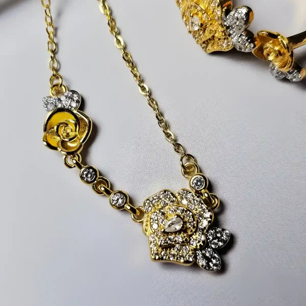 Schmuckset mit Halskette und Damenring in Rosen Design Nahaufnahme einer goldfarbenen Geschenke Schmuckset-Halskette und Damenring. Der Set zeigt zwei stilisierte Rosenblüten: Eine Blüte ist glatt und poliert, die andere ist dicht mit kleinen, funkelnden Kristallen (Pavé-Optik) besetzt und hat einen größeren Kristall in der Mitte. Die Kette verbindet die Elemente mit kleinen, runden Kristallsteinen.