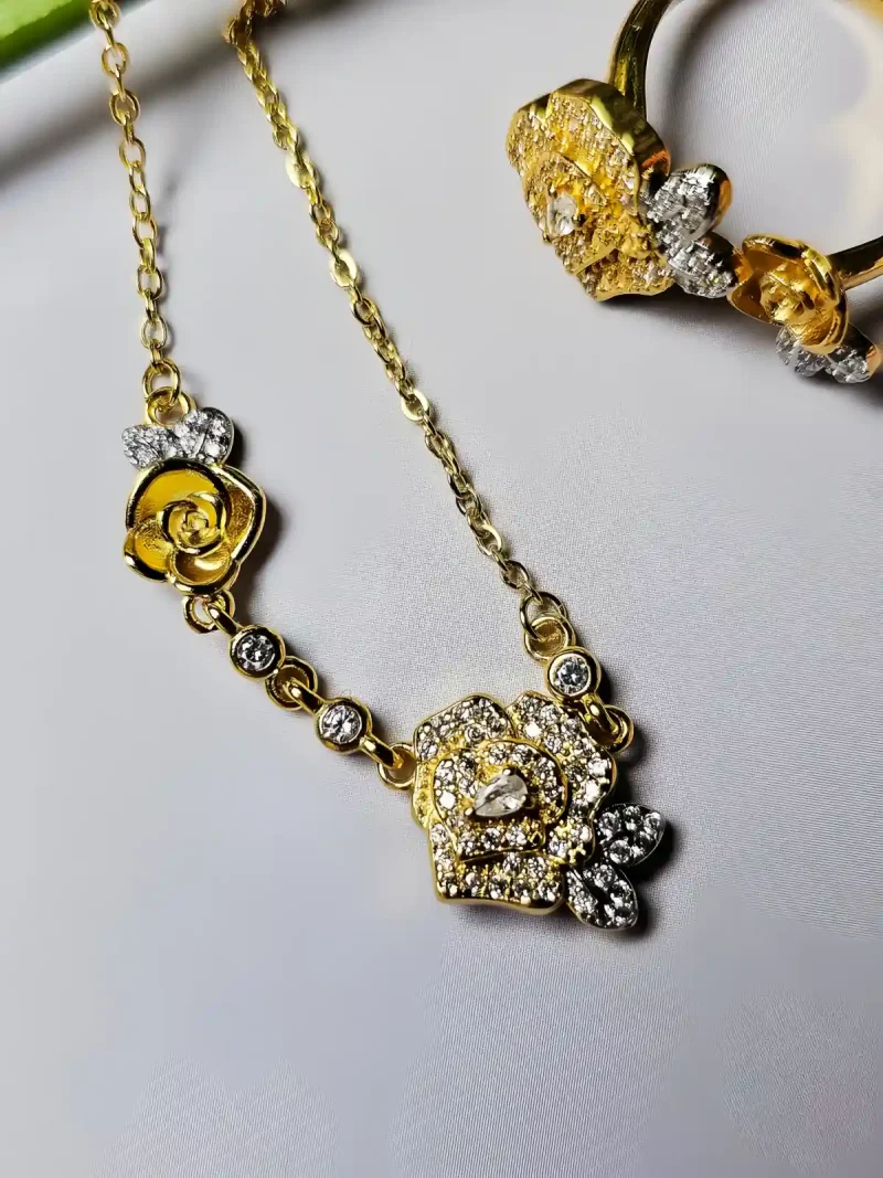 Schmuckset mit Halskette und Damenring in Rosen Design Nahaufnahme einer goldfarbenen Geschenke Schmuckset-Halskette und Damenring. Der Set zeigt zwei stilisierte Rosenblüten: Eine Blüte ist glatt und poliert, die andere ist dicht mit kleinen, funkelnden Kristallen (Pavé-Optik) besetzt und hat einen größeren Kristall in der Mitte. Die Kette verbindet die Elemente mit kleinen, runden Kristallsteinen.