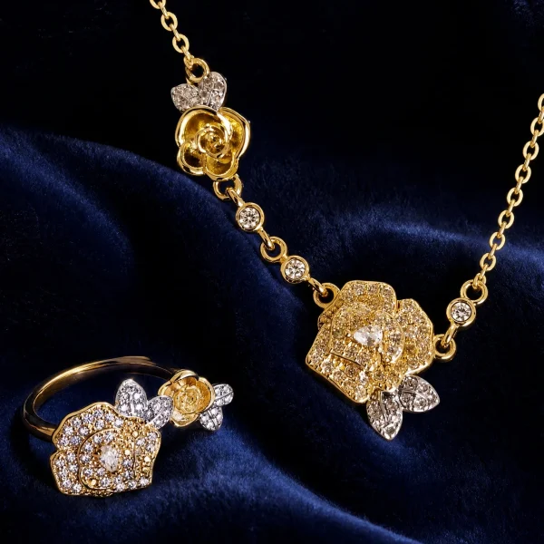 Nahaufnahme einer goldfarbenen Schmuckset-Halskette und Damenring. Der Set zeigt zwei stilisierte Rosenblüten: Eine Blüte ist glatt und poliert, die andere ist dicht mit kleinen, funkelnden Kristallen (Pavé-Optik) besetzt und hat einen größeren Kristall in der Mitte. Die Kette verbindet die Elemente mit kleinen, runden Kristallsteinen.