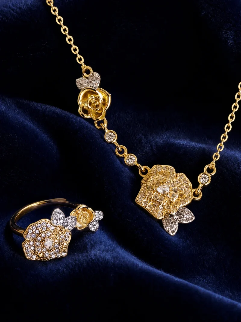 Nahaufnahme einer goldfarbenen Schmuckset-Halskette und Damenring. Der Set zeigt zwei stilisierte Rosenblüten: Eine Blüte ist glatt und poliert, die andere ist dicht mit kleinen, funkelnden Kristallen (Pavé-Optik) besetzt und hat einen größeren Kristall in der Mitte. Die Kette verbindet die Elemente mit kleinen, runden Kristallsteinen.