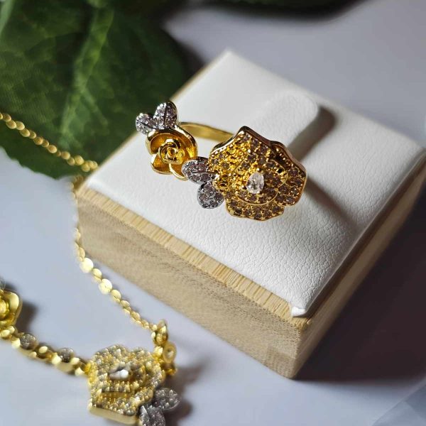 SET-TDG-01-05 Nahaufnahme einer goldfarbenen Schmuckset-Halskette und Damenring. Der Set zeigt zwei stilisierte Rosenblüten: Eine Blüte ist glatt und poliert, die andere ist dicht mit kleinen, funkelnden Kristallen (Pavé-Optik) besetzt und hat einen größeren Kristall in der Mitte. Die Kette verbindet die Elemente mit kleinen, runden Kristallsteinen.
