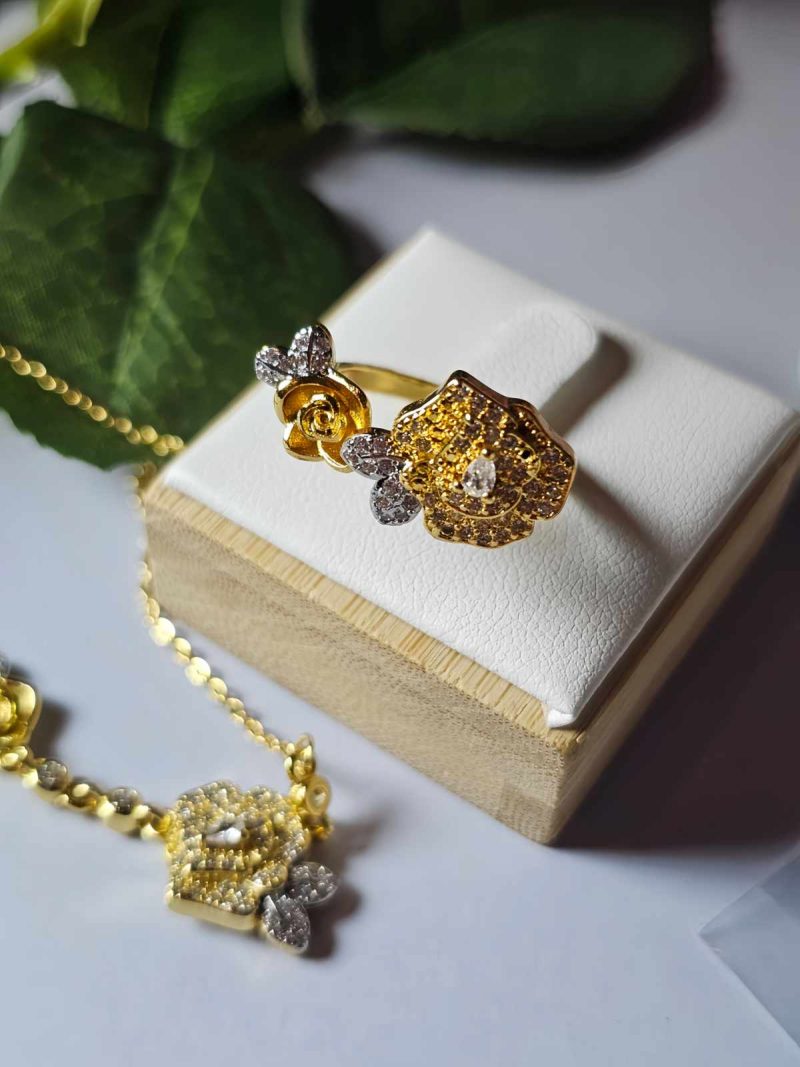SET-TDG-01-05 Nahaufnahme einer goldfarbenen Schmuckset-Halskette und Damenring. Der Set zeigt zwei stilisierte Rosenblüten: Eine Blüte ist glatt und poliert, die andere ist dicht mit kleinen, funkelnden Kristallen (Pavé-Optik) besetzt und hat einen größeren Kristall in der Mitte. Die Kette verbindet die Elemente mit kleinen, runden Kristallsteinen.