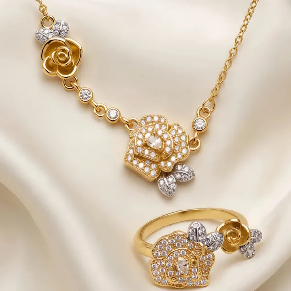 Nahaufnahme einer goldfarbenen Geschenke Schmuckset-Halskette. Der Set zeigt zwei stilisierte Rosenblüten: Eine Blüte ist glatt und poliert, die andere ist dicht mit kleinen, funkelnden Kristallen (Pavé-Optik) besetzt und hat einen größeren Kristall in der Mitte. Die Kette verbindet die Elemente mit kleinen, runden Kristallsteinen.