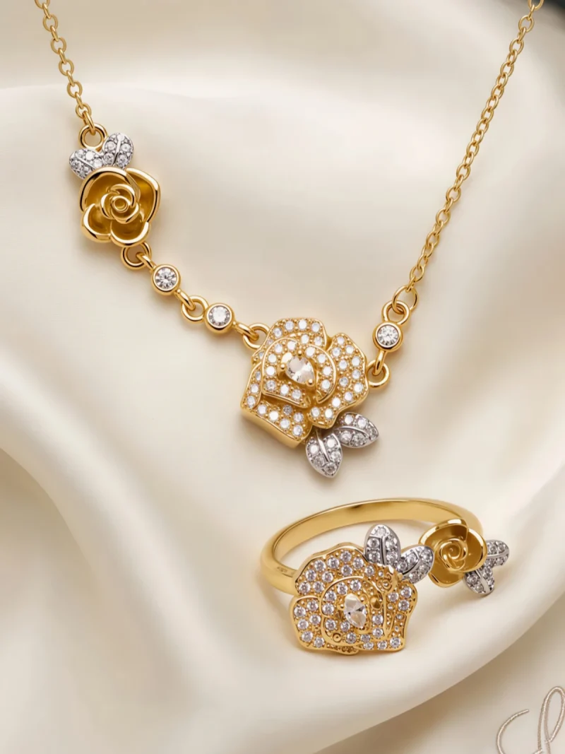 Nahaufnahme einer goldfarbenen Geschenke Schmuckset-Halskette. Der Set zeigt zwei stilisierte Rosenblüten: Eine Blüte ist glatt und poliert, die andere ist dicht mit kleinen, funkelnden Kristallen (Pavé-Optik) besetzt und hat einen größeren Kristall in der Mitte. Die Kette verbindet die Elemente mit kleinen, runden Kristallsteinen.