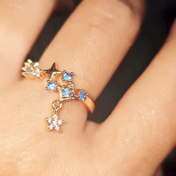 Ein Hand trägt einen goldener, offener Ring mit einem verspielten Design aus Sternen und blauen sowie klaren Zirkonia-Steinen, von dem ein kleiner, mit Steinen besetzter Sternanhänger herabhängt.