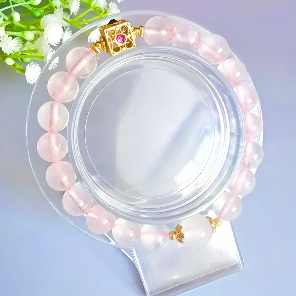 Damen Armband Rosenquarz Armband aus rosafarbenem Quarz mit 8 mm glatten Perlen, verziert mit einem quadratischen, goldfarbenen Verschluss mit Rubin und Onyx. Präsentiert auf transparentem Acrylständer neben zarten weißen Gipsblumen für eine weibliche, elegante Inszenierung.