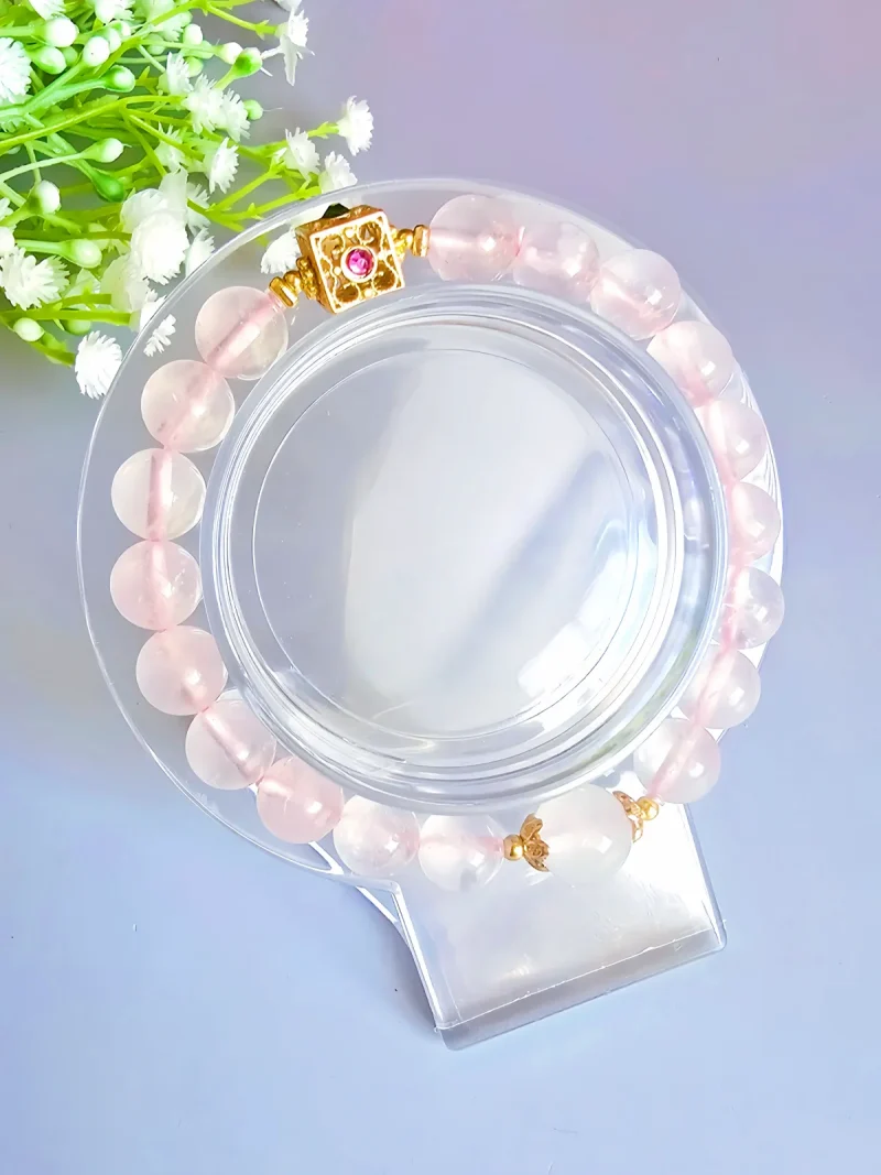 Damen Armband Rosenquarz Armband aus rosafarbenem Quarz mit 8 mm glatten Perlen, verziert mit einem quadratischen, goldfarbenen Verschluss mit Rubin und Onyx. Präsentiert auf transparentem Acrylständer neben zarten weißen Gipsblumen für eine weibliche, elegante Inszenierung.