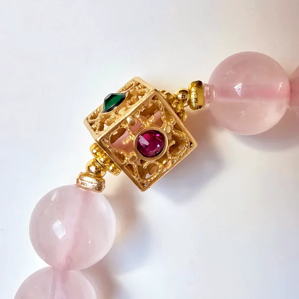 Damen Armband Rosenquarz mit goldfarbenen filigranem Details Nahaufnahme des dekorativen, goldfarbenen quadratischen Verschlusses eines Rosakristall-Armbands: filigrane Durchbrucharbeit, zentrale runde rote und seitliche grüne Zirkonia-Einlagen, 8 mm glatte Perlen im Hintergrund.