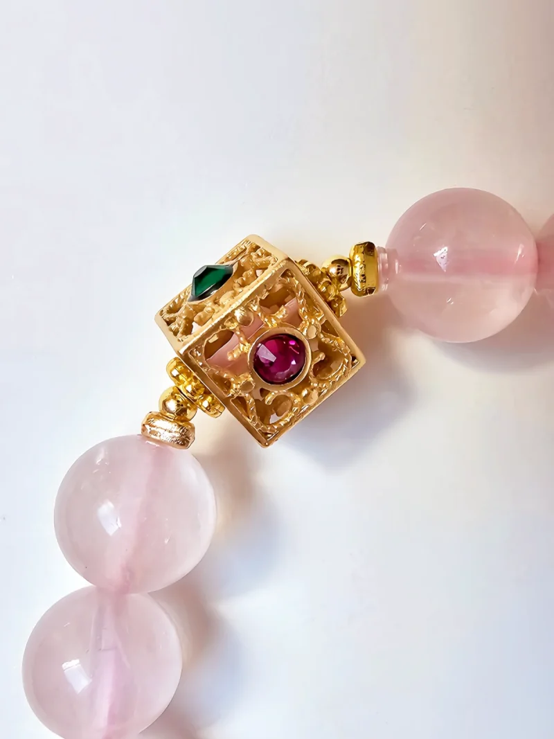 Damen Armband Rosenquarz mit goldfarbenen filigranem Details Nahaufnahme des dekorativen, goldfarbenen quadratischen Verschlusses eines Rosakristall-Armbands: filigrane Durchbrucharbeit, zentrale runde rote und seitliche grüne Zirkonia-Einlagen, 8 mm glatte Perlen im Hintergrund.