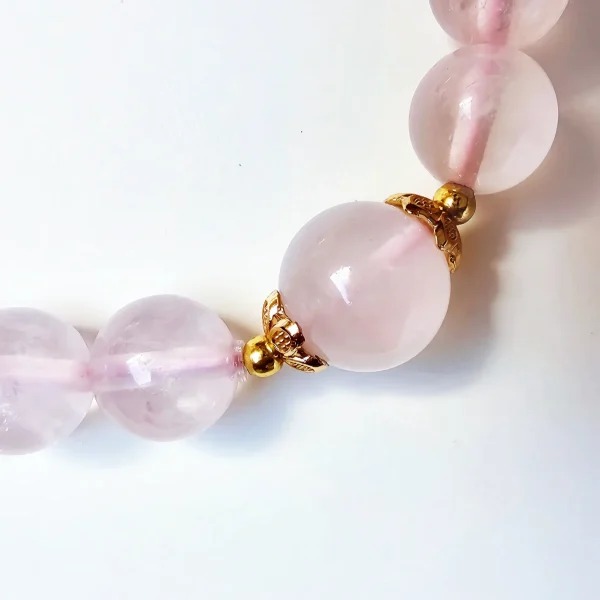 Damen Armband Rosenquarz mit goldfarbenen filigranem Details Detailaufnahme eines Rosakristall-Armbands: glatte, 8 mm runde Perlen aus natürlichem Rosakristall mit sanfter Rosa-Färbung und inneren Einschlüssen, getrennt durch goldfarbene, blütenförmige Metalltrenner. Auf weißem Hintergrund, weiches Licht.