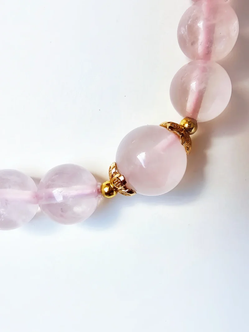 Damen Armband Rosenquarz mit goldfarbenen filigranem Details Detailaufnahme eines Rosakristall-Armbands: glatte, 8 mm runde Perlen aus natürlichem Rosakristall mit sanfter Rosa-Färbung und inneren Einschlüssen, getrennt durch goldfarbene, blütenförmige Metalltrenner. Auf weißem Hintergrund, weiches Licht.