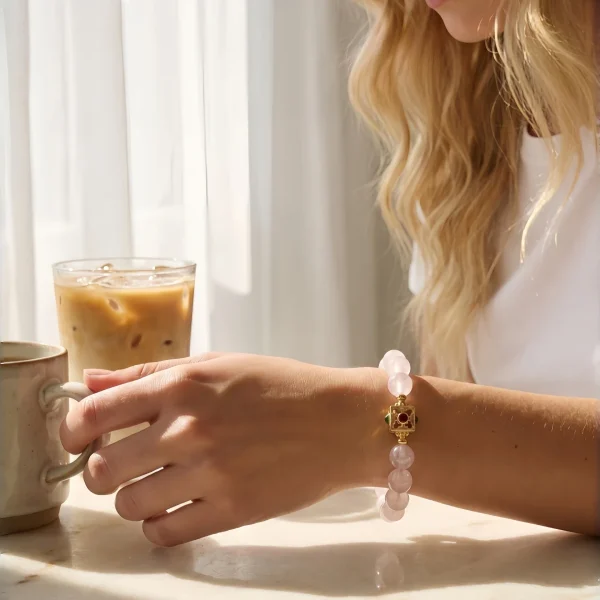 Quarz Armband Perlenarmband Naturstein Eine junge Frau mit blonden Locken trägt ein rosafarbenes Quarz-Armband mit 8 mm glatten Perlen und einem dekorativen, goldfarbenen quadratischen Verschluss mit Rubin- und Onyx-Einlagen. Sie sitzt an einem Marmortisch bei Sonnenlicht, hält eine Keramiktasse und neben ihr steht ein Glas Eiskaffee – eine entspannte, alltägliche Szene, die das Armband als zeitloses Accessoire im Daily Wear zeigt.
