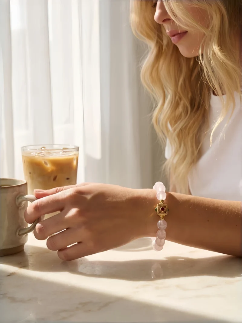 Quarz Armband Perlenarmband Naturstein Eine junge Frau mit blonden Locken trägt ein rosafarbenes Quarz-Armband mit 8 mm glatten Perlen und einem dekorativen, goldfarbenen quadratischen Verschluss mit Rubin- und Onyx-Einlagen. Sie sitzt an einem Marmortisch bei Sonnenlicht, hält eine Keramiktasse und neben ihr steht ein Glas Eiskaffee – eine entspannte, alltägliche Szene, die das Armband als zeitloses Accessoire im Daily Wear zeigt.