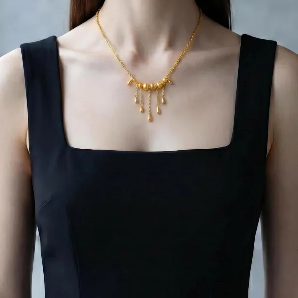 Elegante Goldene Tropfen-Fransen Halskette – Vintage Laser-Design mit schimmernden Kugeln Porträt einer Junge Frau mit dunkle Schwarz Hair. Sie trägt eine Schwarz U-schnitt ohne Arme kurz Shirt und eine filigrane, goldfarbene Halskette. Der Anhänger der Kette besteht aus einer horizontalen Reihe kleiner, glänzender Kugeln, von denen mehrere feine Kettchen mit Tropfen-Formen herabhängen (Vintage-Fransen-Design).