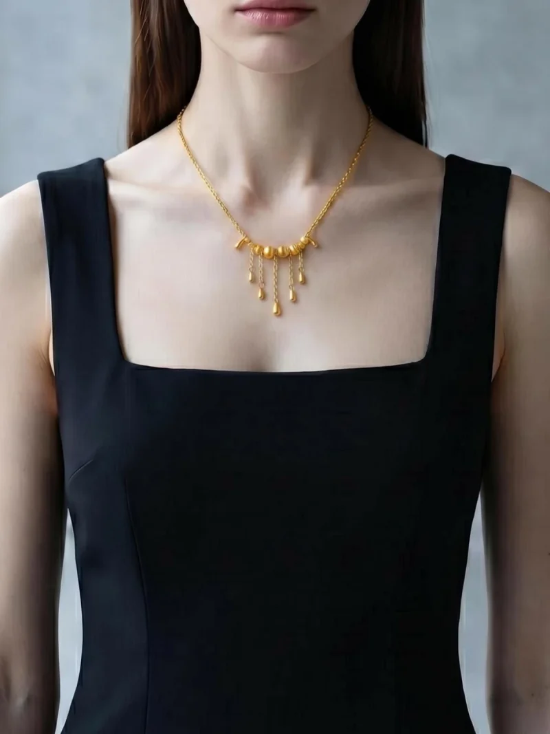 Elegante Goldene Tropfen-Fransen Halskette – Vintage Laser-Design mit schimmernden Kugeln Porträt einer Junge Frau mit dunkle Schwarz Hair. Sie trägt eine Schwarz U-schnitt ohne Arme kurz Shirt und eine filigrane, goldfarbene Halskette. Der Anhänger der Kette besteht aus einer horizontalen Reihe kleiner, glänzender Kugeln, von denen mehrere feine Kettchen mit Tropfen-Formen herabhängen (Vintage-Fransen-Design).