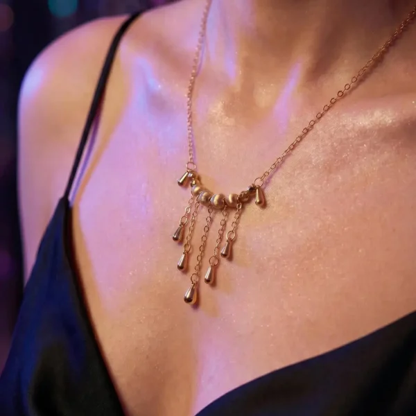 Elegante Goldene Tropfen-Fransen Halskette – Vintage Laser-Design mit schimmernden Kugeln Porträt einer Frau am Abend, Sie trägt eine Schwarz V-Ausschnitt Kleidung und eine filigrane, goldfarbene Halskette. Der Anhänger der Kette besteht aus einer horizontalen Reihe kleiner, glänzender Kugeln, von denen mehrere feine Kettchen mit Tropfen-Formen herabhängen (Vintage-Fransen-Design).