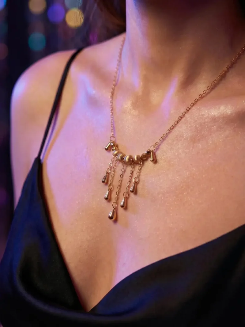Elegante Goldene Tropfen-Fransen Halskette – Vintage Laser-Design mit schimmernden Kugeln Porträt einer Frau am Abend, Sie trägt eine Schwarz V-Ausschnitt Kleidung und eine filigrane, goldfarbene Halskette. Der Anhänger der Kette besteht aus einer horizontalen Reihe kleiner, glänzender Kugeln, von denen mehrere feine Kettchen mit Tropfen-Formen herabhängen (Vintage-Fransen-Design).