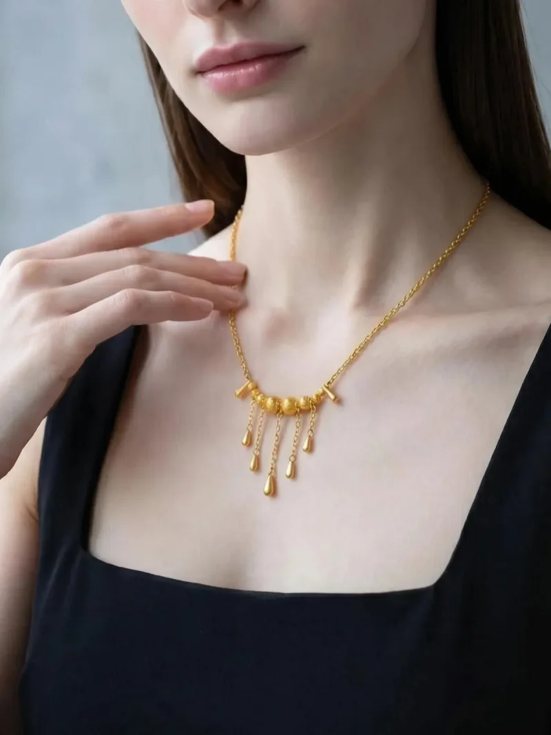 Elegante Goldene Tropfen-Fransen Halskette – Vintage Laser-Design mit schimmernden Kugeln Porträt einer Junge Frau mit dunkle Schwarz Hair. Sie trägt eine Schwarz U-schnitt T-Shirt und eine filigrane, goldfarbene Halskette. Der Anhänger der Kette besteht aus einer horizontalen Reihe kleiner, glänzender Kugeln, von denen mehrere feine Kettchen mit Tropfen-Formen herabhängen (Vintage-Fransen-Design).