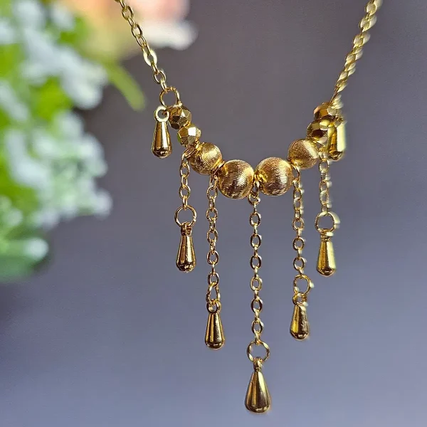 Elegante Goldene Tropfen-Fransen Halskette – Vintage Laser-Design mit schimmernden Kugeln Goldfarbene Modeschmuck-Halskette aus Kupfer, bestehend aus einer feinen Kette und einem mittigen Anhänger mit drei geriffelten Laser-Kugeln, von denen fünf filigrane Ketten mit kleinen Tropfen-Anhängern herabfallen.