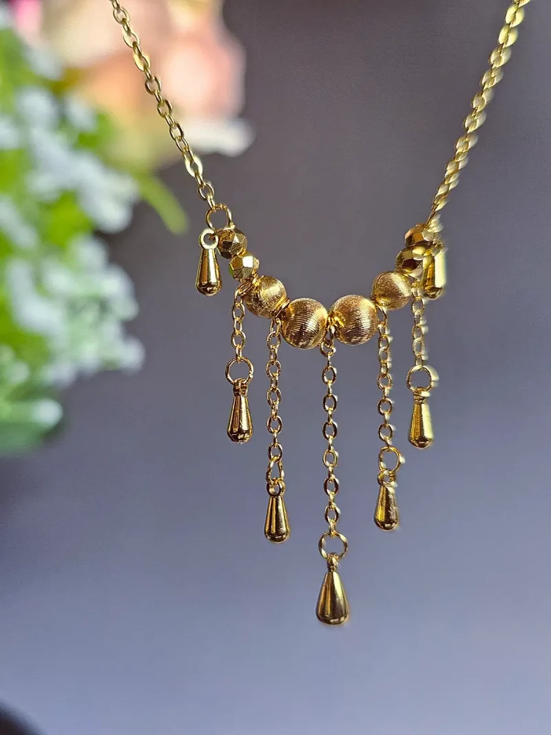 Elegante Goldene Tropfen-Fransen Halskette – Vintage Laser-Design mit schimmernden Kugeln Goldfarbene Modeschmuck-Halskette aus Kupfer, bestehend aus einer feinen Kette und einem mittigen Anhänger mit drei geriffelten Laser-Kugeln, von denen fünf filigrane Ketten mit kleinen Tropfen-Anhängern herabfallen.